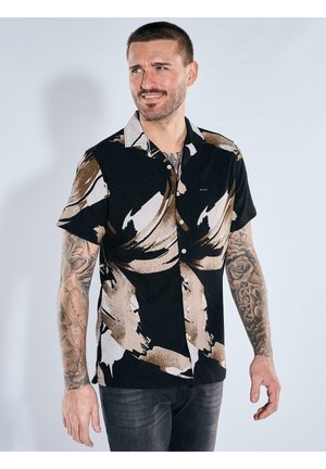 Mann mit kurzen Haaren und Tattoos trägt ein schwarzes kurzärmeliges Hemd mit beigen abstrakten Mustern, schaut zur Seite und lächelt.