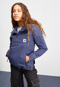 Marineblaue Windjacke mit einer Reißverschluss-Brusttasche, verstellbaren Manschetten und Seitentaschen; mit einem Logopatch auf der Vorderseite.