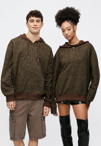 Sudadera con capucha con estampado de leopardo marrón, cremallera frontal, capucha y puños marrones, con un ajuste holgado. Un modelo lleva shorts cargo grises, y otro lleva shorts negros.