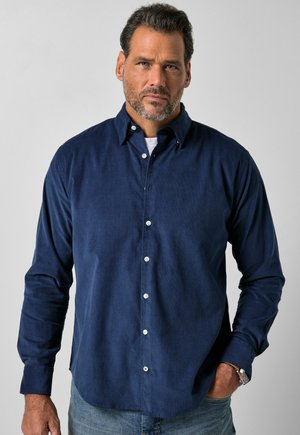 Mann mit kurzen dunklen Haaren und Bart, trägt ein dunkelblaues Hemd mit Knöpfen und hellblaue Jeans, mit einer Hand in der Tasche vor einfarbigem Hintergrund.