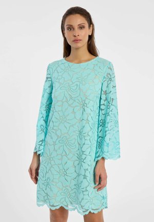 Femme portant une robe en dentelle florale bleu clair à manches longues et ourlet festonné, debout devant un fond blanc uni.