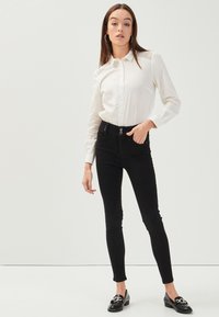 Camicia bianca con bottoni, maniche lunghe, abbinata a jeans neri slim-fit e mocassini neri. Tessuto liscio e design aderente con bottoni visibili.