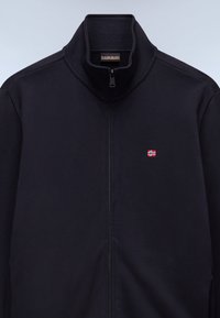 Μαύρο μπουφάν με φερμουάρ σε όλο το μήκος και γιακά που στέκεται όρθιος, με ένα μικρό κόκκινο και λευκό έμβλημα σημαίας στο αριστερό στήθος και ορατή ετικέτα μάρκας στο εσωτερικό του γιακά.