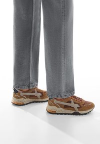 Zapatillas marrones y beige con parte superior texturizada, cordones grises y una suela media moteada; combinadas con pantalones vaqueros grises holgados.