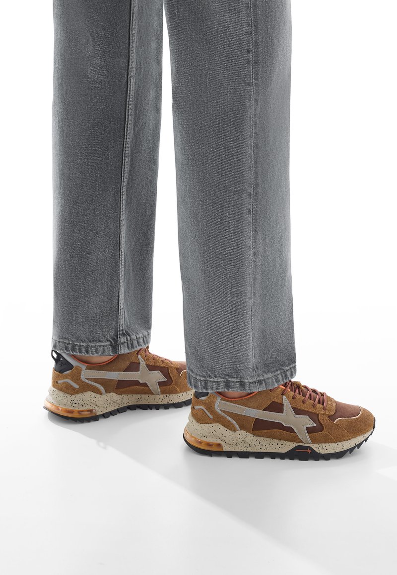 Zapatillas marrones y beige con parte superior texturizada, cordones grises y una suela media moteada; combinadas con pantalones vaqueros grises holgados.