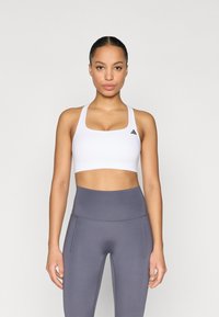 Fehér sportmelltartó négyzet alakú nyakkivágással és széles pántokkal, fekete logóval díszítve. Szürke, magasderekú leggingsszel van párosítva, oldalsó varrásokkal.