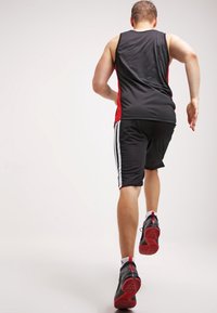 Débardeur et short de sport noir et rouge, avec des rayures blanches sur les côtés. Le coureur porte des chaussures noires avec des semelles rouges. Vue arrière.