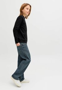 Fiatal személy áll, fekete hosszú ujjú inget, bő kék farmert és fehér tornacipőt visel, egyik kezével zsebében.