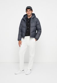 Uomo con cappello nero, giacca a vento scura sopra una camicia a quadri, pantaloni bianchi e sneakers bianche, in piedi con le mani in tasca su uno sfondo bianco.