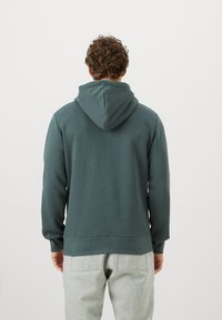 Görbe hajú férfi, sötétzöld kapucnis pulóverben és világosszürke melegítőnadrágban áll, háttal egy sima háttér előtt.