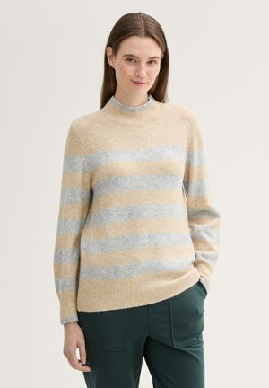 Gestreifter Strickpullover mit weichen beigen und hellgrauen horizontalen Streifen, hochribbeligem Kragen und ribbeligen Bündchen. Kombiniert mit einer dunkelgrünen Hose.