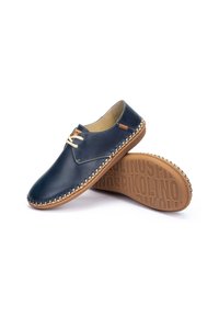 Zapatos de cuero azul marino con punta redonda, cordones beige, costuras decorativas y una suela marrón texturizada con un patrón de logotipo de la marca.