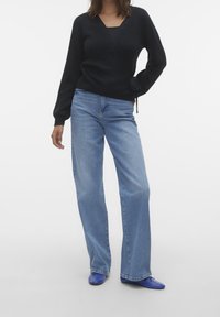 Svart omslagströja med långa ärmar, kombinerad med ljusblå vida jeans. Skorna är blå, vilket ger en avslappnad, lager-på-lager look.