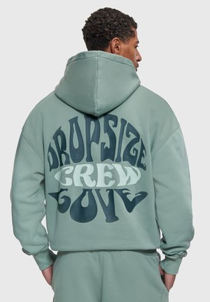 Homme portant un sweat à capuche surdimensionné vert clair et un pantalon assorti, dos affichant un grand texte incurvé "DROPSIZE CREW GLOVE" en bleu foncé et blanc.