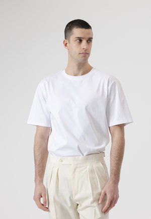 Joven con cabello corto y oscuro que lleva una camiseta blanca lisa metida dentro de pantalones crema plisados de cintura alta, mirando hacia un lado.