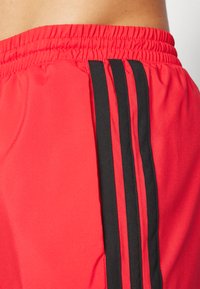 Red athletic shorts with a textured surface, featuring three vertical black stripes along the side and an elastic waistband.