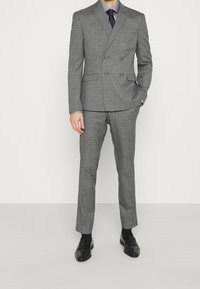 Abito doppiopetto a quadri grigi con rever a lancia, due tasche frontali, vestibilità sartoriale, abbinato a una cravatta scura e scarpe nere lucide.