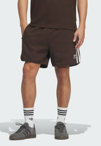 Hnědé ribbed šortky se zipy po stranách, které mají tři bílé pruhy podél boků a malé logo Adidas na spodní přední části.