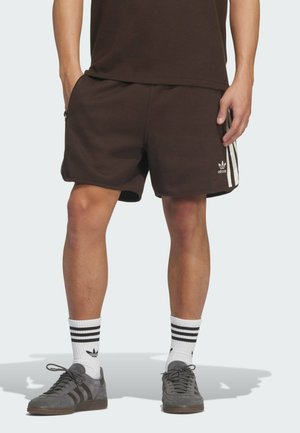 Shorts marroni a coste con zip laterali, caratterizzati da tre strisce bianche lungo i lati e un piccolo logo Adidas sulla parte anteriore inferiore.