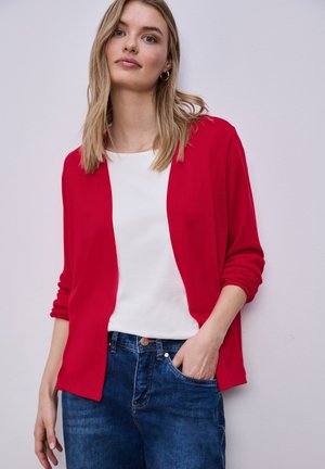 Jeune femme portant un cardigan rouge ouvert sur un haut blanc, un jean bleu, une main dans la poche, sur un fond clair uni.