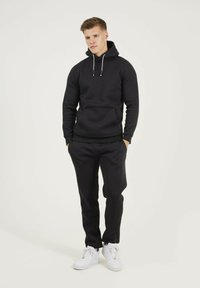 Haut à capuche noir avec une poche kangourou à l'avant et un cordon de serrage. Associé à un pantalon noir et des baskets blanches. Texture de tissu lisse.