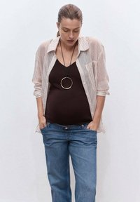 Mujer vestida con una camiseta a rayas sobre una blusa negra y jeans azules, con las manos en los bolsillos, la cabeza inclinada hacia abajo, frente a un fondo blanco liso.