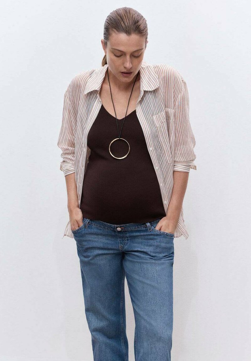 Mujer vestida con una camiseta a rayas sobre una blusa negra y jeans azules, con las manos en los bolsillos, la cabeza inclinada hacia abajo, frente a un fondo blanco liso.