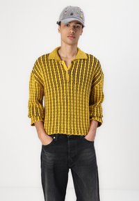 Gul- og sort stribet strikpullover med krave og knaplukning, parret med sorte jeans. Grå kasket med broderede detaljer.