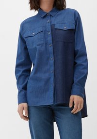 Kvinde iført en langærmet, knap-op denimskjorte med to brystlommer, kombineret med blå jeans.
