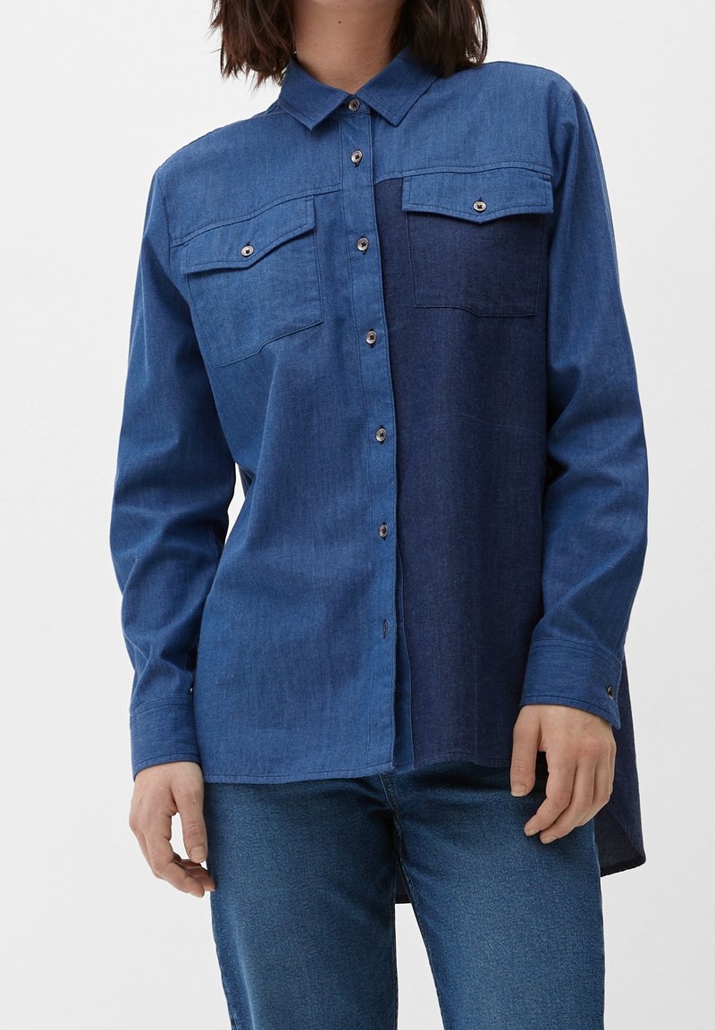 Kvinde iført en langærmet, knap-op denimskjorte med to brystlommer, kombineret med blå jeans.