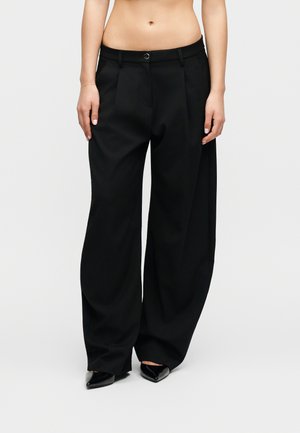 Άτομο που φοράει ψηλόμεσα, φαρδιά μαύρα παντελόνια με πιέτες μπροστά και μαύρα παπούτσια με μυτερό μύτη, στέκεται μπροστά σε απλό φόντο.