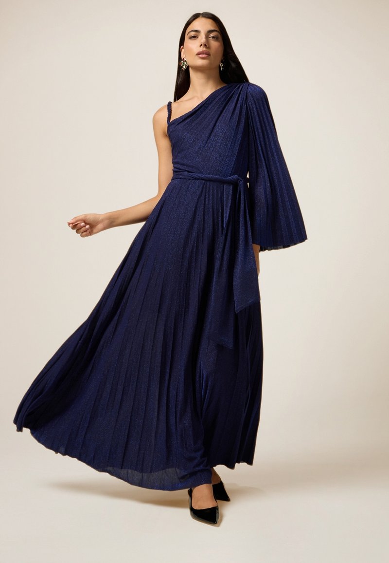 Navy blue, laskinettu iltapuku yhdellä olkapäällä, epäsymmetrinen hiha ja sidottu vyötärö. Sileä pinta ja hienovarainen hohde. Mustat korot jalassa.