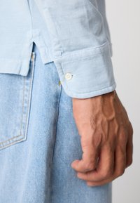 Mannens hånd ved siden av en lyseblå denimjeanslomme, iført en lyseblå langermet skjorte med knappeleddet.