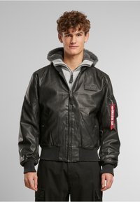 Veste aviateur en cuir noir avec une capuche doublée en fausse fourrure grise, des poignets et un ourlet côtelés. Comprend une fermeture éclair et une poche latérale avec une étiquette.