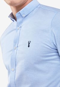 Lichtblauw overhemden gemaakt van zachte stof, met een klein zwart geborduurd hertlogo op de borst en een standaard kraag.