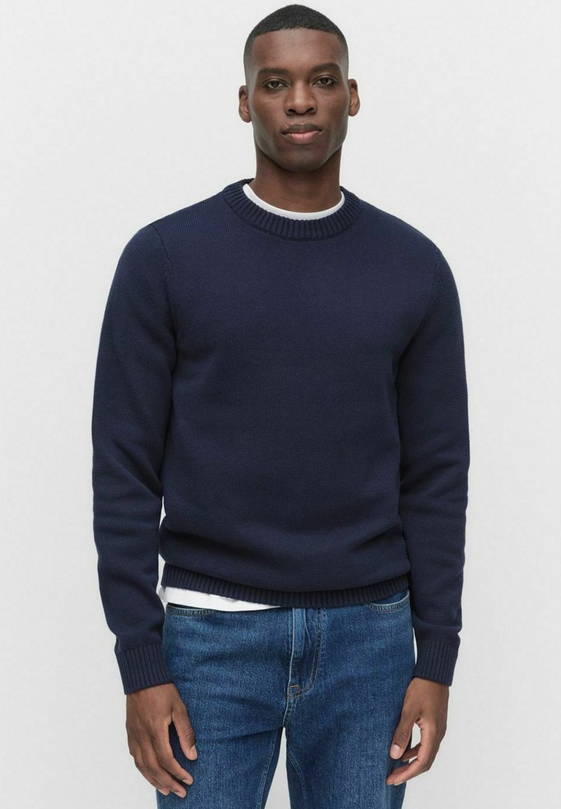 Marinblå stickad tröja med rundad halsringning, ribbade ärmslut och nederkant; bärs över en vit T-shirt, ihop med blå denimjeans.