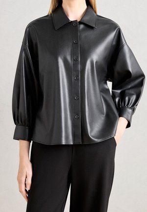 Vrouw draagt een zwarte nep leren blouse met kraag en driekwart ballonmouwen, gecombineerd met een zwarte broek.