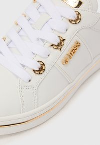 Λευκό αθλητικό παπούτσι με λευκά κορδόνια, χρυσά διακοσμητικά οπές, χρυσό λογότυπο "GUESS" στο πλάι και χρυσή επένδυση στη σόλα.