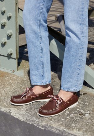 Brązowe skórzane buty typu boat shoes z białymi podeszwami i sznurówkami, noszone z jasnoniebieskimi jeansami na betonowych schodach w pobliżu metalowej konstrukcji.