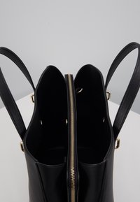 Sac fourre-tout en cuir noir avec deux compartiments principaux ouverts et une section centrale zippée, montrant des détails de fermeture éclair et de ferrures dorées.