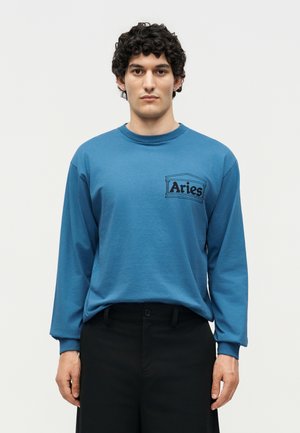 Mand med kort krøllet hår iført blå langærmet skjorte med "Aries"-logo på brystet og sorte bukser, der står mod en ensfarvet baggrund.