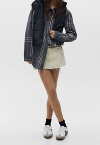 Veste matelassée noire portée sur une chemise à carreaux gris et blanc, associée à une jupe en denim beige avec un ourlet effiloché. Baskets blanches avec des accents gris.