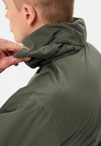 Groene, lichtgewicht jas met een hoge kraag. De stof heeft een gestructureerd oppervlak. Een hand past de kraag aan, waardoor details zichtbaar worden.