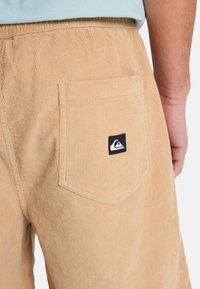 Παντελόνια κοτλέ σε ανοιχτό καφέ χρώμα· διαθέτουν πίσω τσέπη με μαύρο λογότυπο και κάθετη ριγέ υφή. Ελαστική μέση για άνεση.