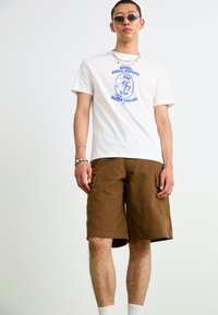 White t-shirt with blue graphic print, paired with brown cargo shorts. Chunky silver chain necklace and sunglasses, white socks visible.