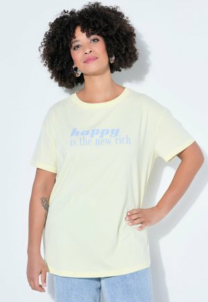 Mujer con cabello rizado, usando pendientes y una camisa amarillo pálido que dice "feliz es el nuevo rico", posando con la mano en la cadera sobre un fondo blanco.