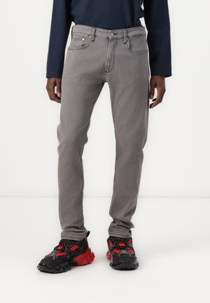 Grå denimjeans med rett snitt, fem-lommers design, kontraststing, og knappelukking. Brukt med svarte og røde sneakers.