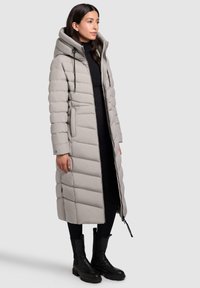 Lange grijze pufferjas met capuchon, voorzien van brede horizontale stiksels, een ritssluiting aan de voorkant en zijzakken. Gestyled met zwarte leggings en laarzen.