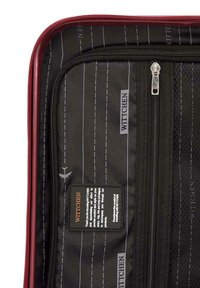Interno di una valigia rossa con zip, foderato in tessuto nero con righe bianche. Include una tasca in rete e un'etichetta del marchio con dettagli testuali.
