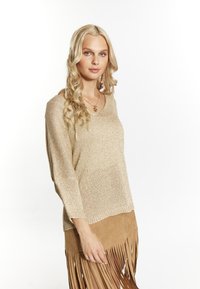 Maglione tricot color oro chiaro con scollo ampio, abbinato a una gonna in suede beige con frange. Il tessuto testurizzato aggiunge interesse visivo.